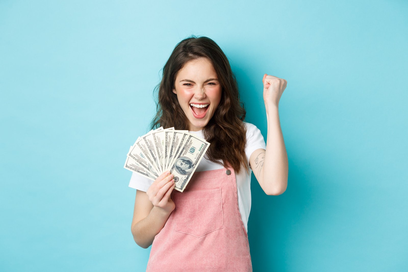 Lucky young woman looks excited, shouting from satisfaction and triumph, winning money, holding dollar bills and making fist pump, standing over blue background Lucky young woman looks excited, shouting from satisfaction and triumph, winning money, holding dollar bills and making fist pump, standing over blue background.